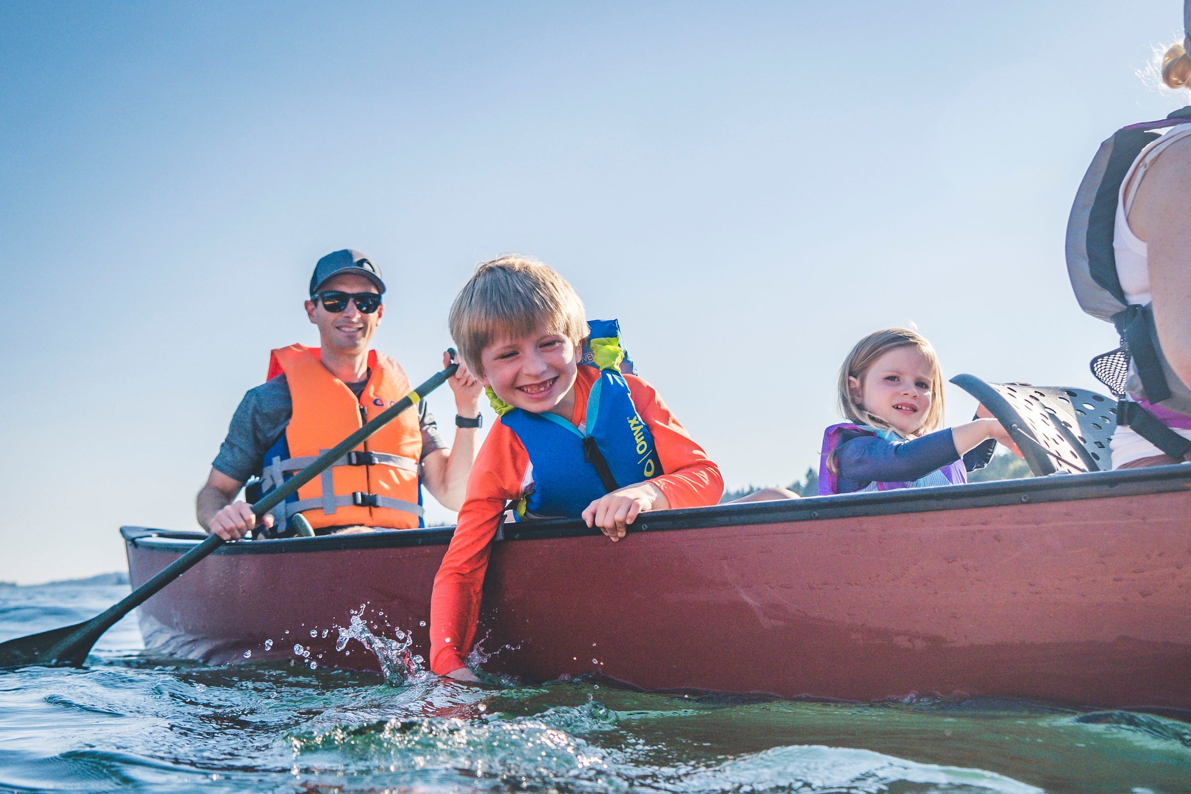 A family smiling while one boy dips his hand into the water from a canoe wearing Onyx Personal Flotation Devices.