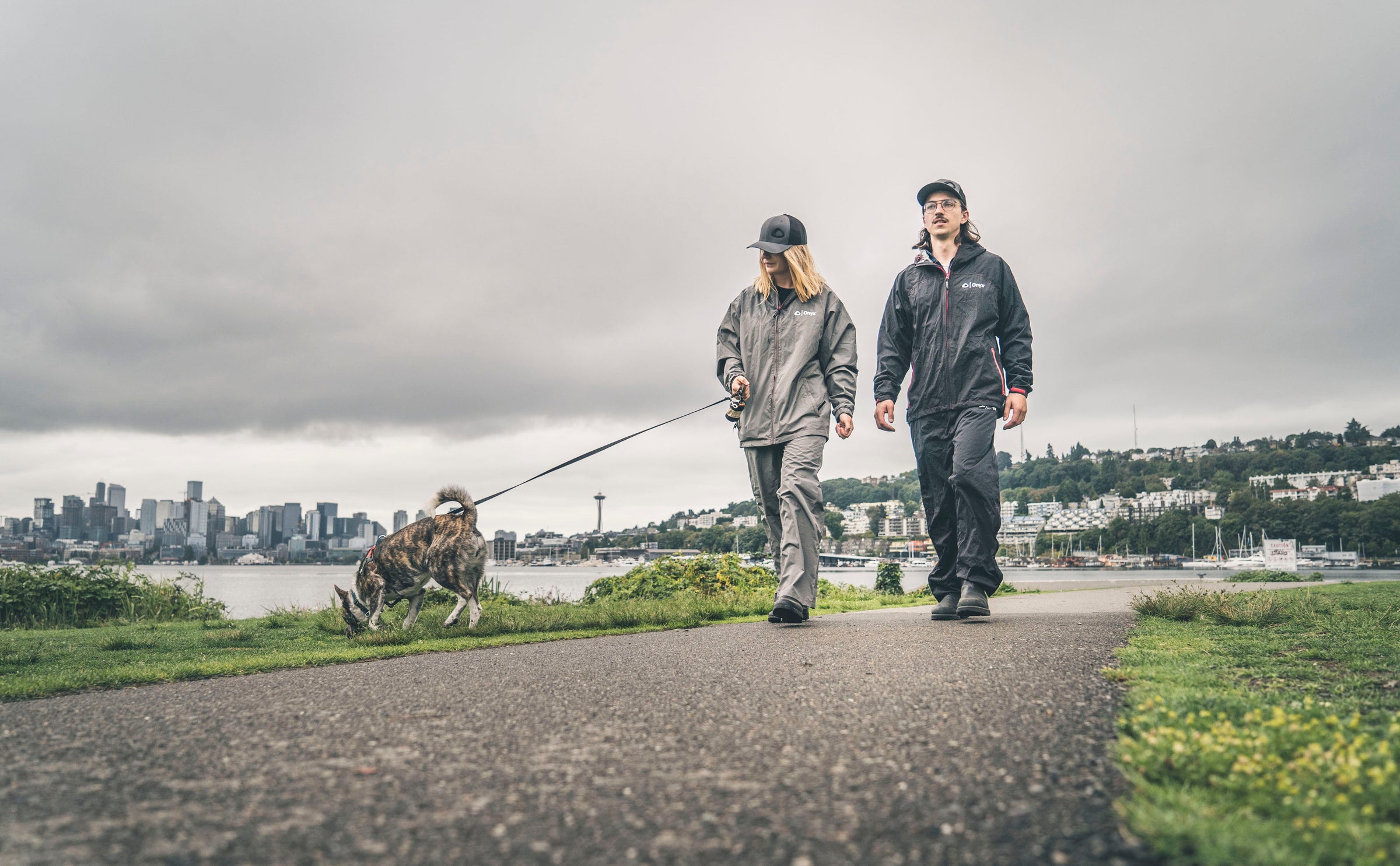 Two people wearing Onyx Apparel walking their dog on a walkway near a body of water.