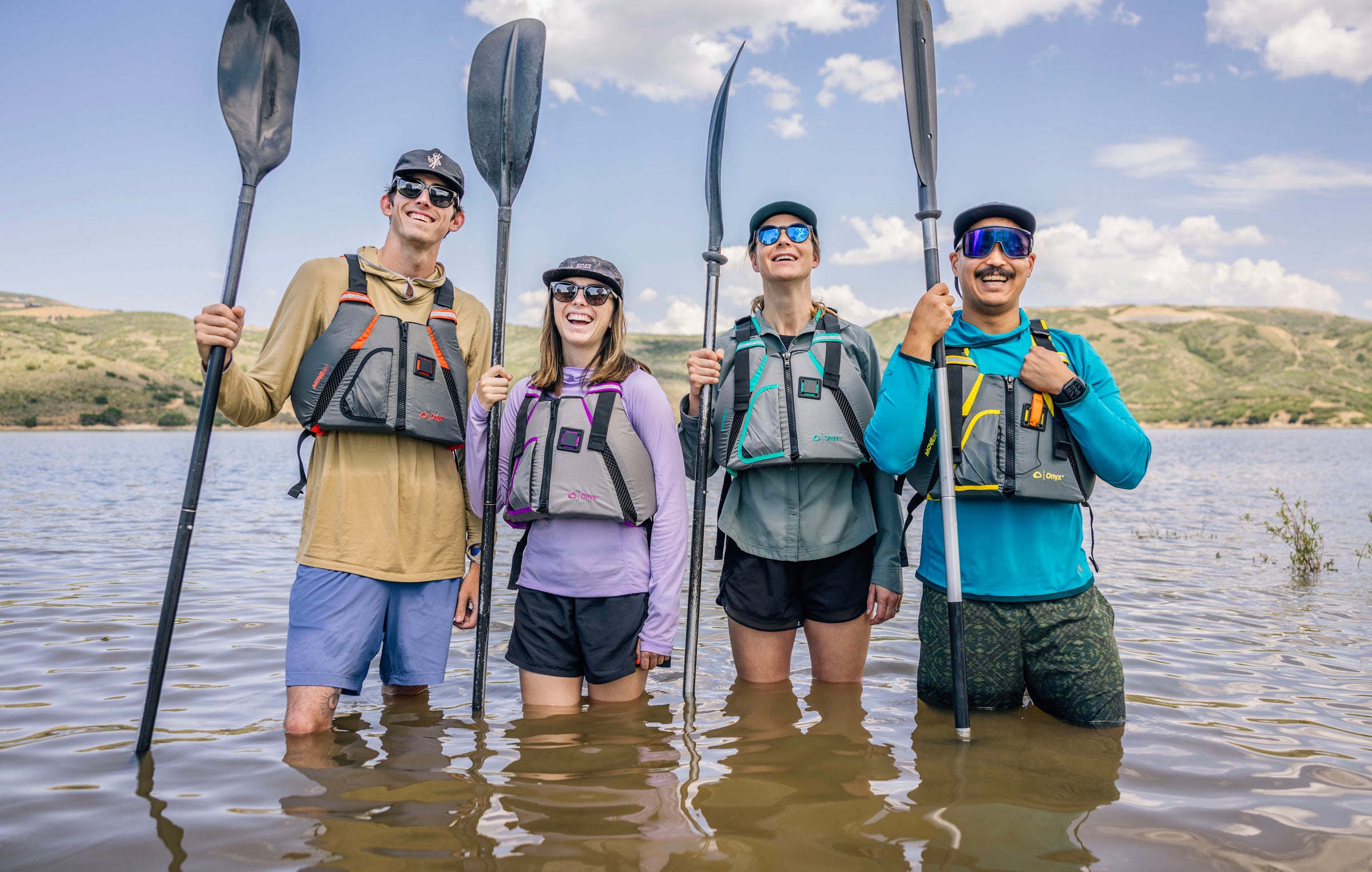 Group of four smiling kayakers standing in lake, wearing Onyx life jackets and holding paddles.
