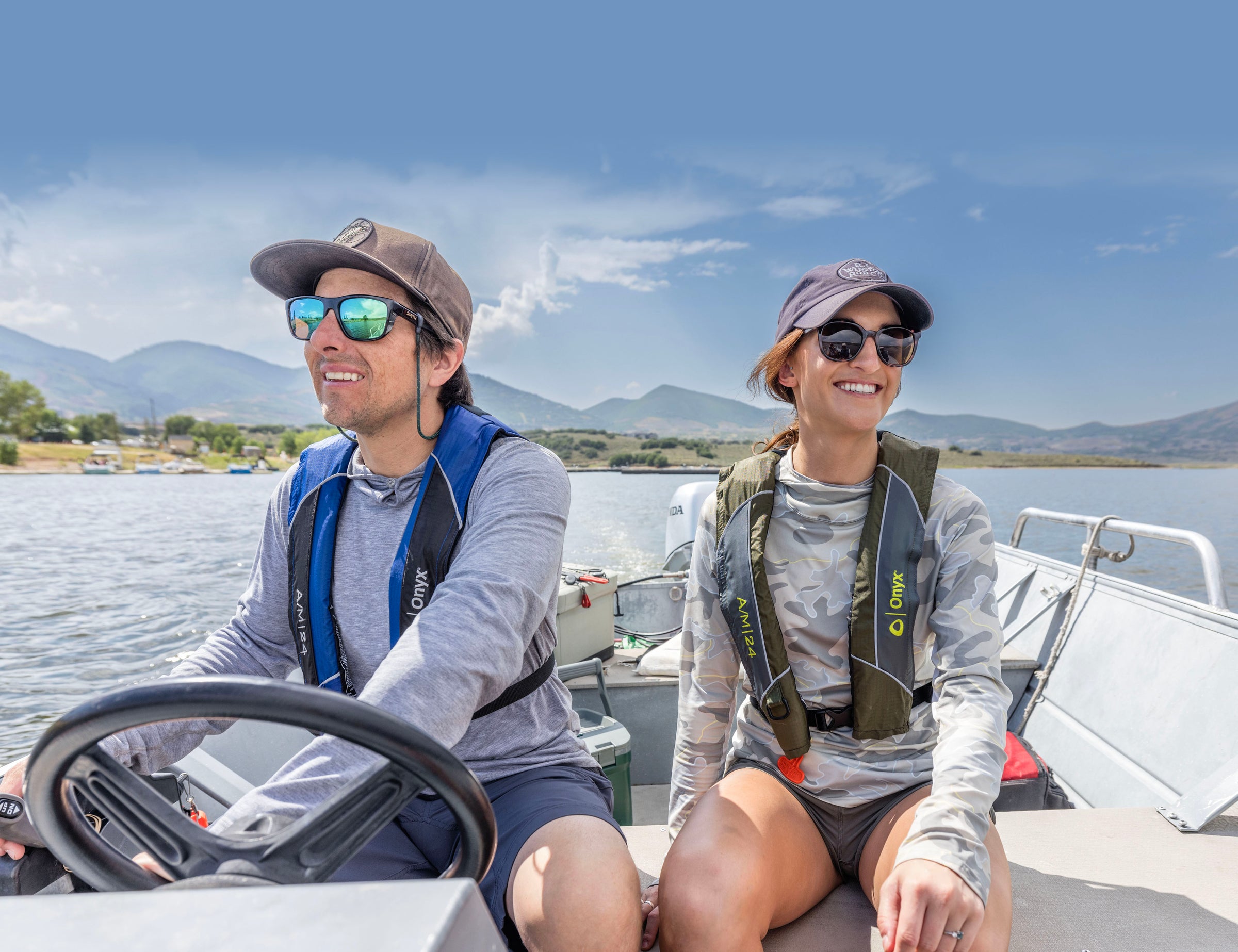 Smiling couple in boat wearing Onyx inflatable life jackets with lake and hills behind.