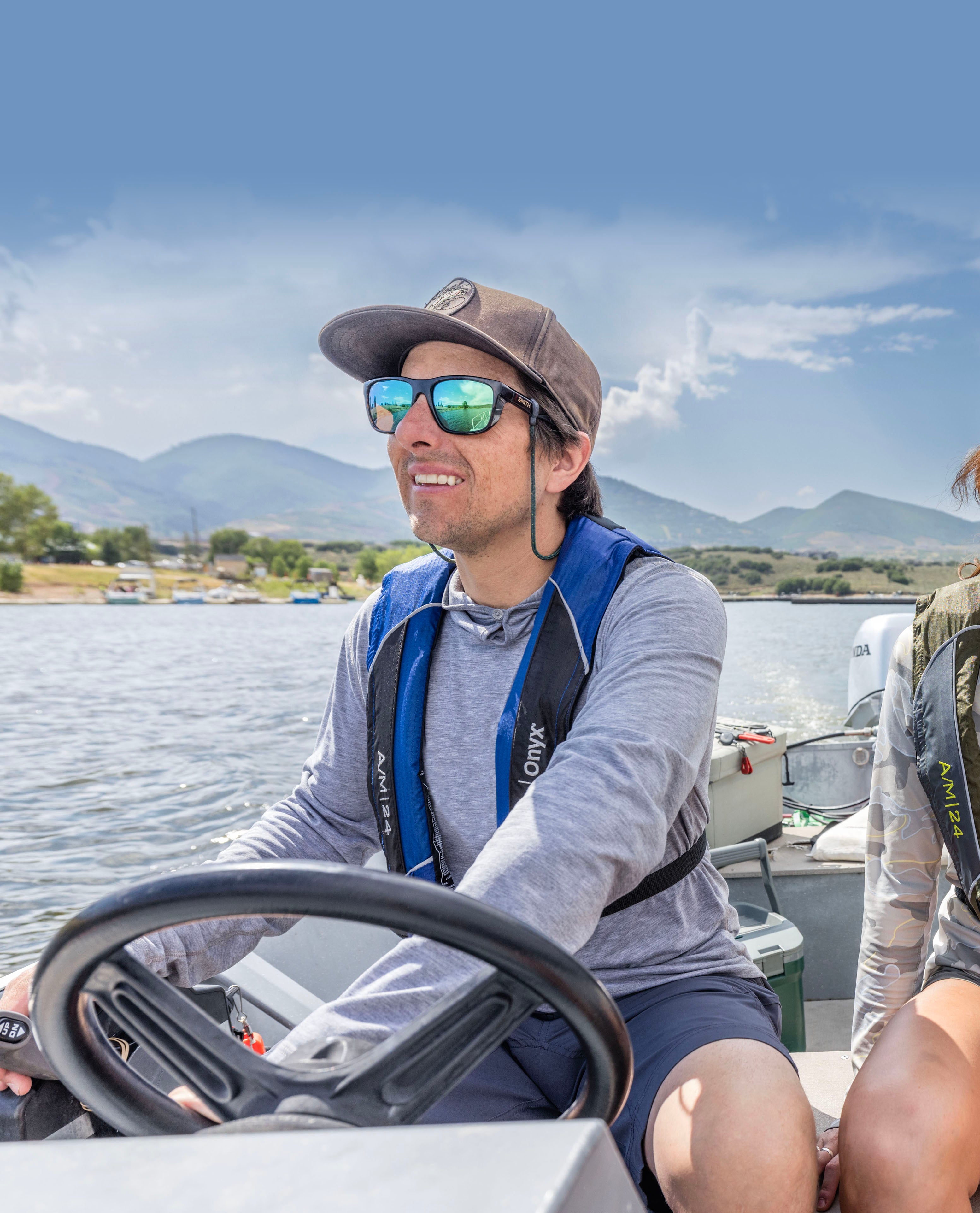 Man steering a boat while wearing an Onyx blue life jacket, with lake and mountains behind.
