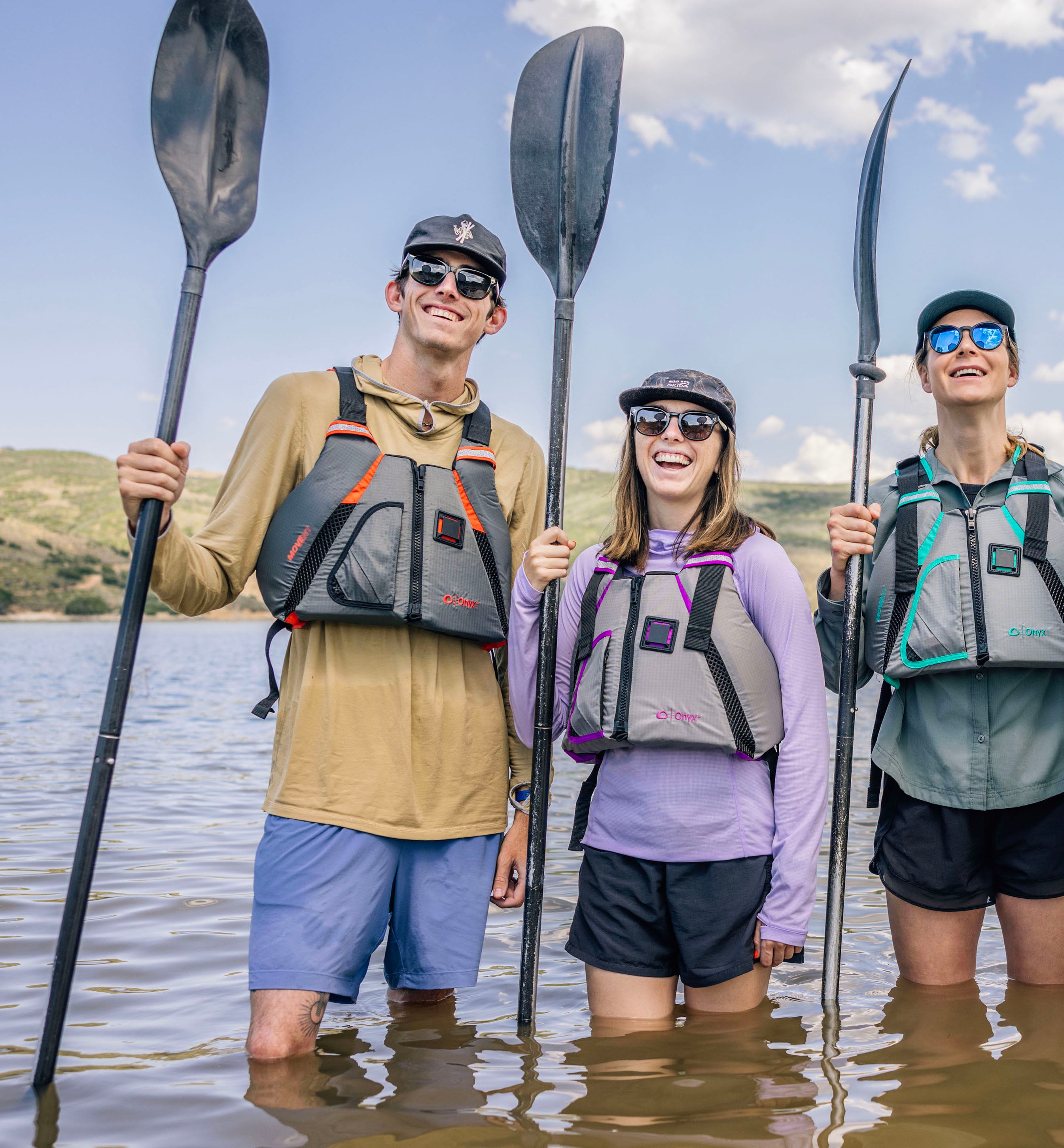 Three smiling people standing in water wearing Onyx life jackets and holding paddles.