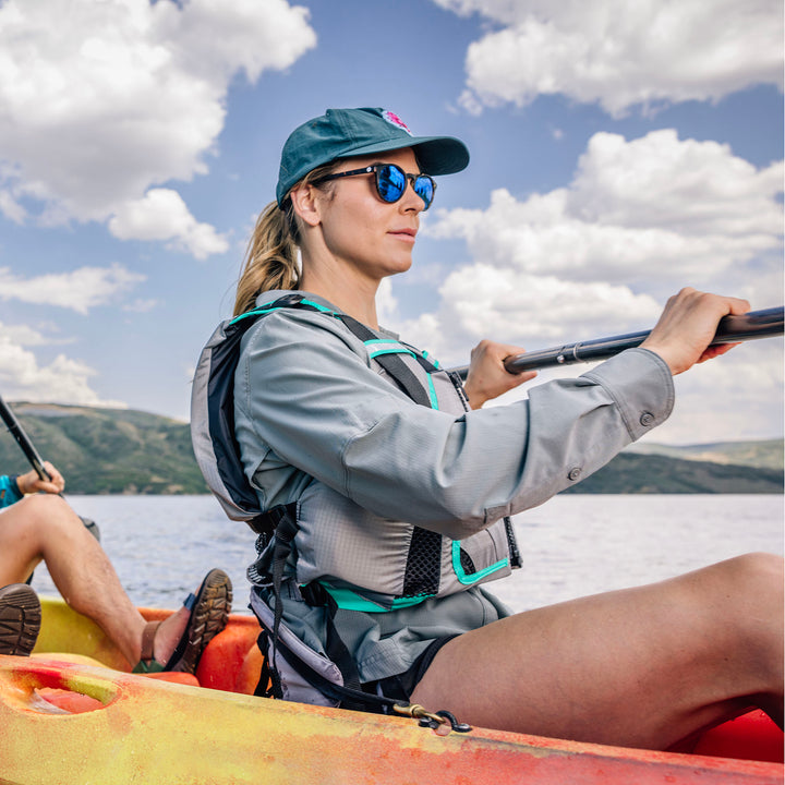 Woman kayaking on a lake, wearing a MoveVent Dynamic Life Jacket, cap, and sunglasses.